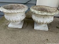 Front and side view of two white concrete planters with square bases, outdoors on a concrete surface, showing paint chipping and general wear.
