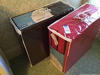 Two Fold-A-Beds in original storage bags, one dark brown and one red, standing upright on carpet.