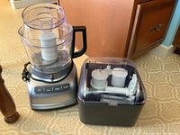 Photo of the KitchenAid food processor base with clear bowl and black handle next to a storage box containing attachments.