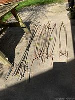 Seven metal garden items laid out on a concrete patio: four slender obelisks with a rusted finish on the right, three taller shepherd's hooks on the left also rusted, under outdoor shade.