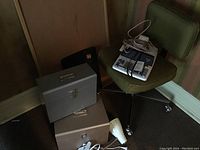 Green fabric desk chair on casters, two metal file boxes stacked, white clip-on desk lamp on floor, brown wall background
