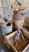 Large rusty milk can, galvanized pail, banding clamp, hand planer and scythe blade arranged on ground.