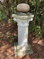 Full view of the concrete Grecian style column standing on ground among trees and pine needles.