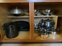 Photo of cabinet contents showing enamelled black speckled tin pots, pans and roasting pans on left side and stainless steel cookware on right