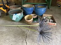 Overview photo showing large ceramic pots, smaller pots, rock collection in box, and spiky metal garden stake lying on concrete floor near wooden wall
