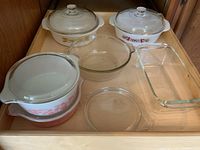 Seven dishes including Fire-King printed casseroles, Pyrex Gooseberry covered casseroles, clear Pyrex casserole dish, spare lid, and loaf pan inside wooden drawer.