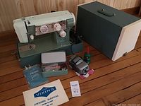 Full view of the sewing machine with carrying case, owner's manual, metal accessory box with bobbins and needles, and oil bottle all displayed on wooden floor.
