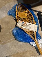 Single Kabir Kouba wooden snowshoe with paper label attached reading 'Kabir Kouba' and featuring an illustration, on blue plastic sheeting.