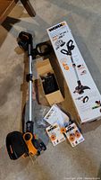 Full view of Worx cordless string trimmer with battery, charger in box, and replacement string spools on concrete floor with original packaging.