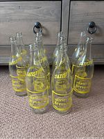 Front view of 10 clear glass Jersey Dry Beverage bottles arranged in a group on carpet in front of wooden drawers.