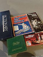 Five vintage board game boxes stacked together showing Pictionary, National Pro Hockey, Super Mastermind, Trivial Pursuit All-Star Sports Edition, and Magnetic Chess and Checker Set.