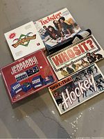 Photo showing all six vintage board games in their original boxes arranged on the floor. Games include Pursue the Pennant, Electric Jeopardy, Strat-O-Matic Hockey, Whosit?, Twister, and Pente.