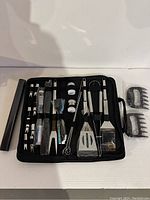 Full set of barbecue tools displayed inside an open black zippered case, including spatula, tongs, fork, skewers, seasoning containers, and specialized utensils.