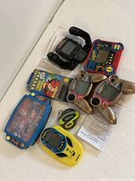 Photo of the full lot of various electronic travel games showing the multiple handheld devices with screens, different game buttons and controls, and some paperwork.
