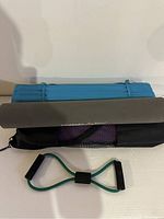 Folded blue exercise mat, grey Life Fitness mat on top, black mesh bag underneath, and green resistance band with foam handles.