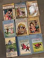 Photo showing 10 paperback Laura Ingalls Wilder novels arranged on a floor surface. The covers depict illustrated scenes and characters from the Little House series. Titles visible include 'Little Town on the Prairie', 'The First Four Years', and others.
