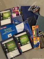 Photo showing mixed office supplies including binders, notebooks, sheet protectors, dividers, pens, correction tape, and file folders spread on a floor.