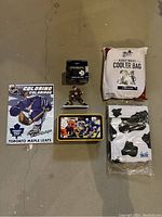 A collection of mixed Toronto Maple Leafs hockey themed items including a coloring book, miniature figurine, collectible tin, a sealed pack of collectible hockey figurines, and a cooler bag.