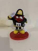 Front view of red M&M fireman dispenser showing character's face, fireman's helmet with yellow 'M', black jacket with yellow stripes, yellow shoes, white gloves, and red base.