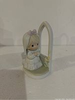 Front view of Precious Moments figurine showing girl in white dress with lavender bow standing beside a white arch with yellow flower details.