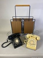 Black and cream vintage rotary telephones with metal and vinyl magazine rack in the background.