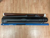 Three fishing rod cases side by side on wood floor, one black Plano AIRLINER, one blue Academy fabric tube, and one brown hard plastic tube