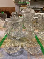 Photo showing two glass ashtrays, pinwheel crystal cookie jar with lid, candy jar with lid, and honey pot, all made of clear glass with intricate cut designs.