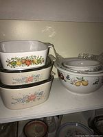 Three stacked casserole dishes with vegetable and floral patterns next to a round bowl with pear motif and white lids.