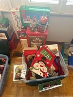 Photo showing plastic storage bin filled with Christmas linens, advent calendar, and various decorations, with boxed Christmas village houses stacked nearby.