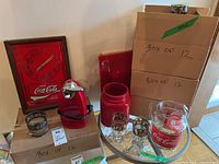 Overall display of Coca-Cola items: wall clock, cookie jar base, stemmed and old-fashioned glasses, pitcher, electric can opener, and labeled boxes.