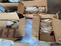 Three open cardboard boxes containing multiple pairs of unused white cotton work gloves and a bundle of white drop cloths on a surface.