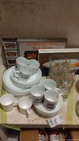 Corelle dishes with floral leaf pattern stacked with matching mugs on top, boxed lacquerware tray set behind, plastic pitcher and glass set visible