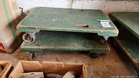 Two weathered green painted wooden dollies stacked showing wheels and size label on side.