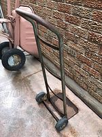 Metal hand dolly leaning against an outdoor wall on a concrete surface. Handle is curved metal with rust, wheels visible on the bottom.