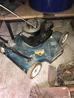 Blue steel deck gas lawn mower on concrete floor with visible white wheels and handle, showing overall condition and construction.
