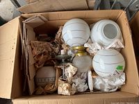 Overhead view of a box containing more than ten vintage glass globe ceiling light fixtures wrapped in protective paper with some brass mounting hardware visible