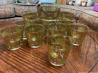 Photo showing twelve rocks glasses with gold and green decorative filigree pattern, ice bucket, and metal tray on wood surface.