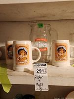 Overall view of set includes one clear glass pitcher with orange and white Hires Root Beer logo and four white ceramic mugs with vintage Hires Root Beer label featuring a child holding a bottle
