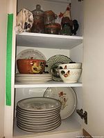 Three shelves inside a cabinet showing various plates, bowls, jars, and a rooster figurine.