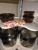 Three large Vision by Corning glass pots with lids and black handles shown stacked on a white corner kitchen shelf, alongside colorful pumpkin themed oven mitts.