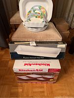 Overview of serving dishes, porcelain rectangular deep plate in box, KitchenAid utensil set box, and colorful floral melamine plates.