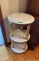 Full view of round three-tier table stand with white marble shelves and gold metal legs near a wooden cabinet.