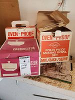 Three boxed items: Connoisseur 2.5 quart covered casserole box in front left, two Cutler oven-proof mixing bowl sets boxes on right, all on a shelf in garage.