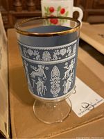 Close-up of a vintage glass with pale blue background, white classical Greek figures, and gold rim.