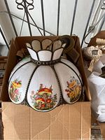 Frontal view of scalloped hanging lamp shade showing white textured glass panels with colored mushroom decals and dark metal framing, inserted in cardboard box.