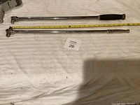 Two breaker bars lying on fabric background with measuring tape showing length, one with rubber grip handle.