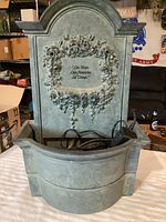 Front view of green cast iron water fountain showing floral design and inscription, basin with electric pump inside.