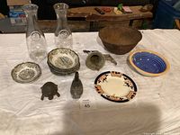 Overview photo of the lot on a white table cover with various china plates, glass carafes, cast iron items, and bowls visible.