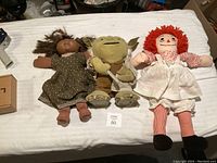 Three dolls lying on a white fabric surface: a brown-skinned doll in a floral dress, a Yoda plush in the middle, and a Raggedy Ann doll on the right.