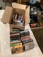 Photo of a cardboard box and stacks of DVDs arranged on a white surface, showing quantity and variety of DVDs in the lot.